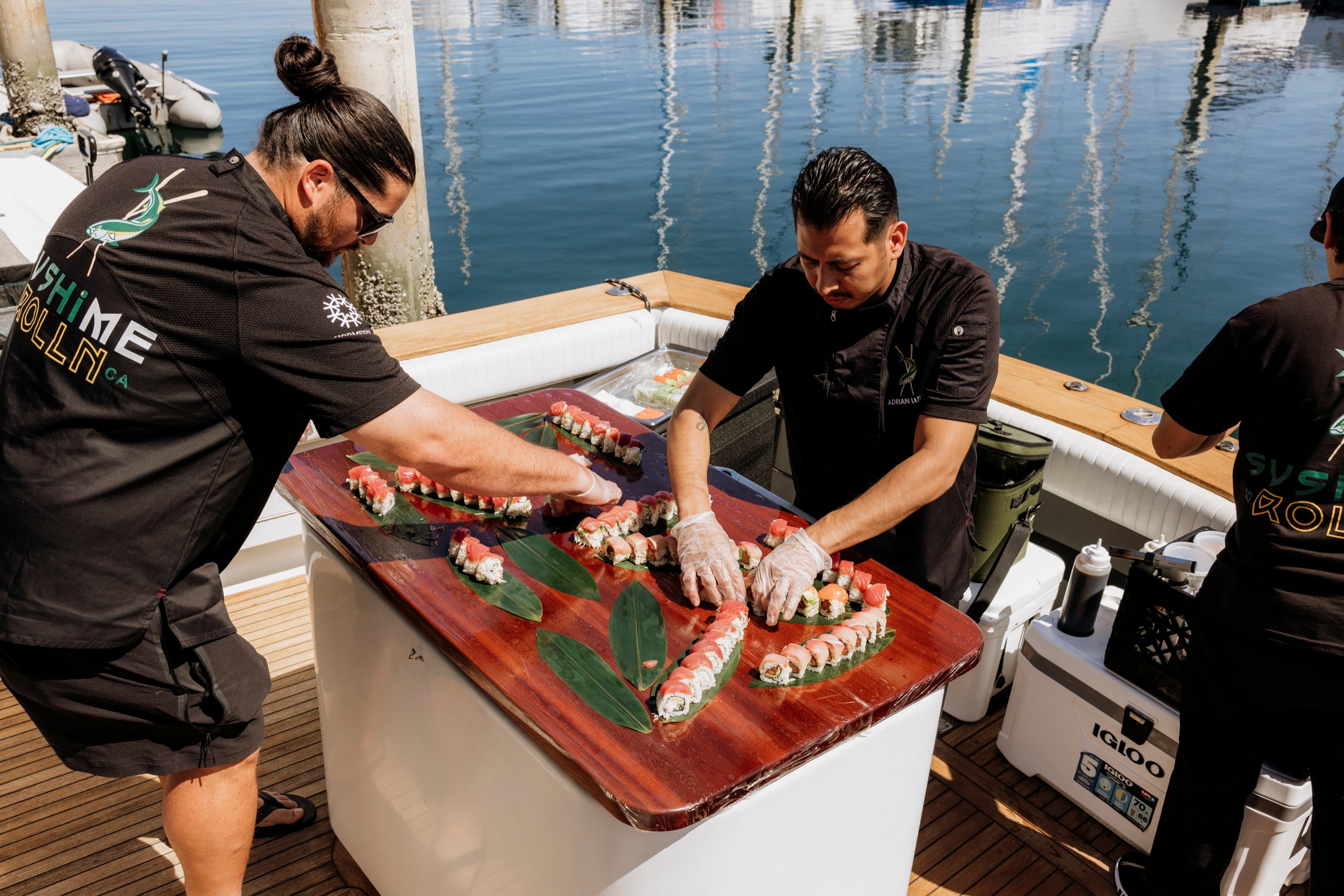 Two sushi chefs plating rolls on a lacquered board on Bluebill's deck with the marina behind